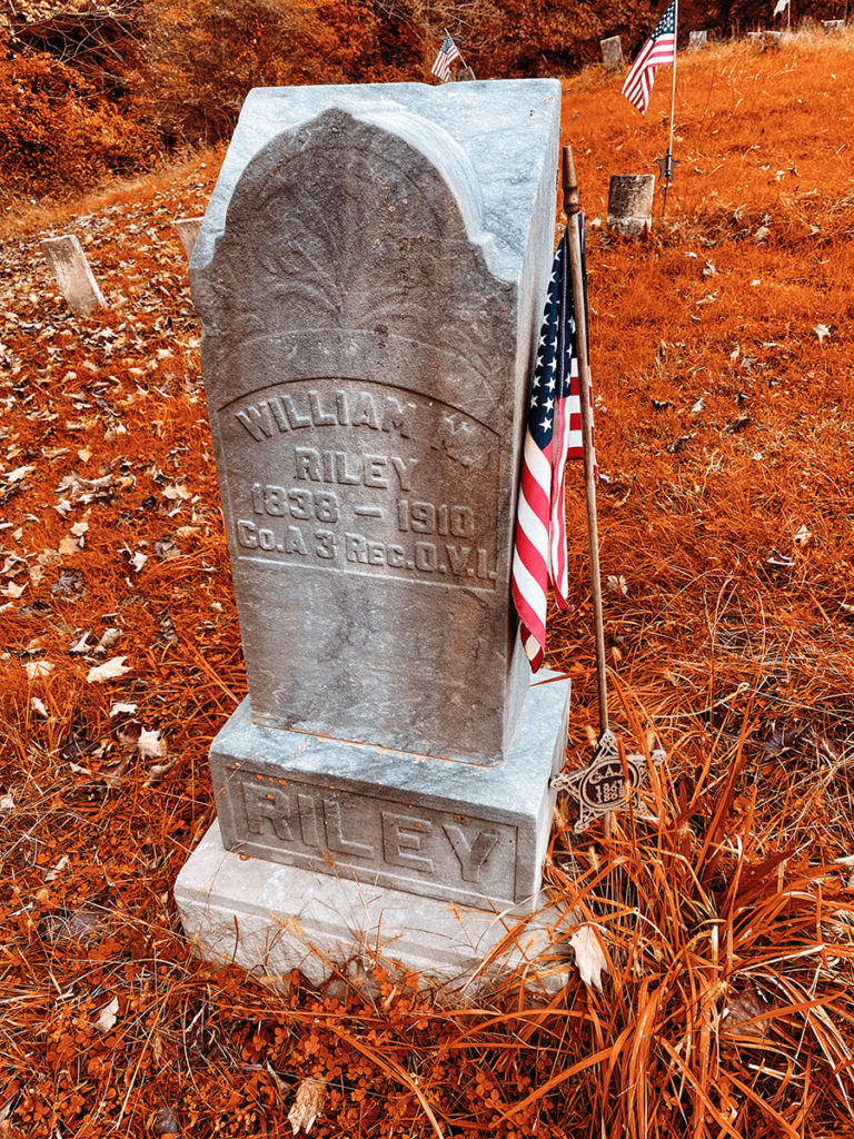 Headstone with an American Flag next to it. Orange fall grass surrounds it.