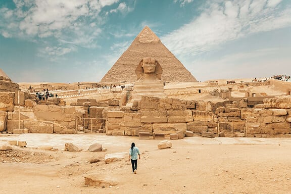 Person standing in the desert in front of Egyptian pyramid.